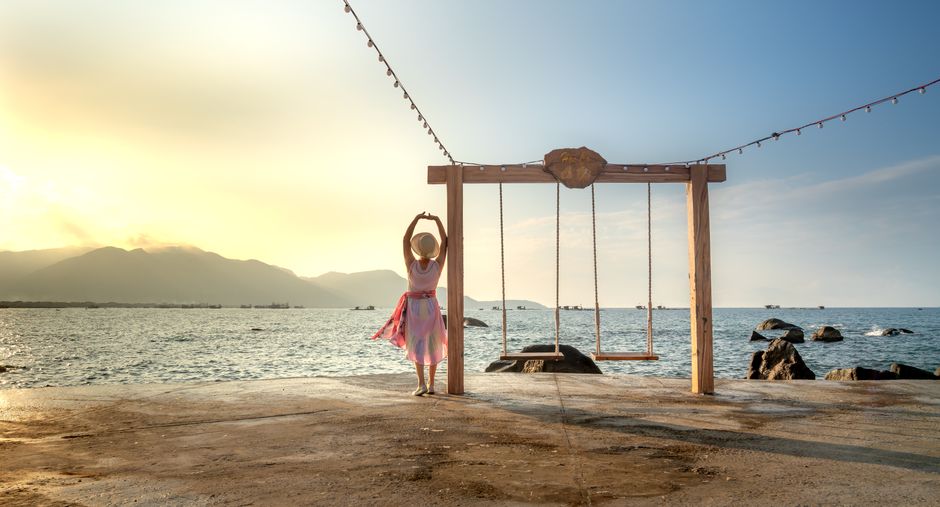 A woman in a hat stands by a swing set, appreciating a beautiful beach sunset.