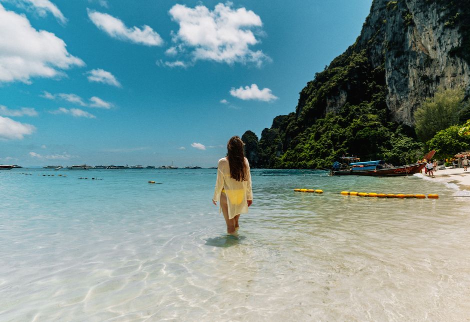 A woman in a yellow dress walks along a picturesque beach in Ko Panyi, Thailand under a clear blue sky.