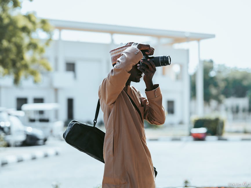 A photographer in traditional attire takes a picture outdoors in a sunny urban setting.