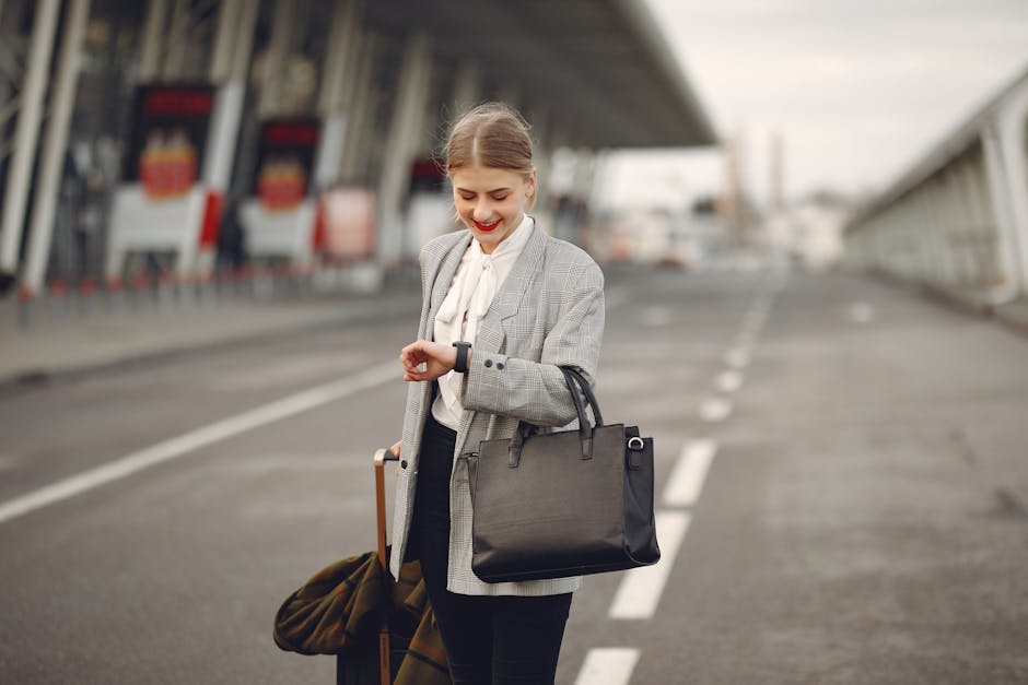 Smiling female passenger in formal outfit checking time on wristwatch while walking along asphalt road to airport terminal with suitcase and handbag during business trip